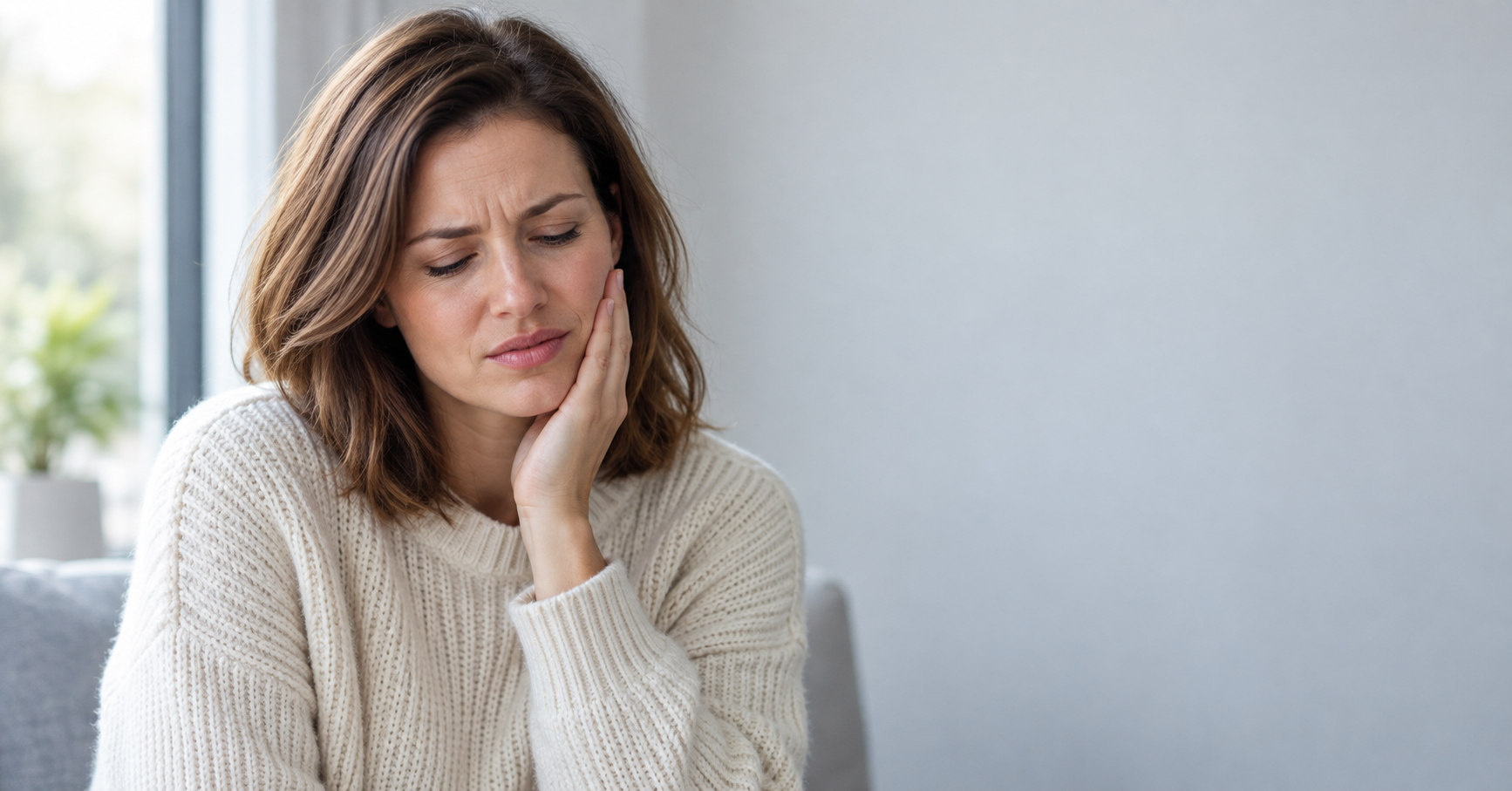 Woman in a cream sweater wincing and holding her cheek, showing why a toothache hurts so much